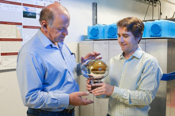 From left, Professor Yuri Efremenko of the University of Tennessee in Knoxville and Jason Newby of Oak Ridge National Laboratory are among 80 participants in COHERENT, a large, collaborative, particle physics experiment to record neutrinos at the Spallation Neutron Source. Photomultiplier tubes look like giant light bulbs and are used to detect light from neutrino interactions in detectors. COHERENTâ€™s cesium iodide detector, the first to espy neutrinos at the SNS, employs a 5-inch (13-centimeter) wide photomultiplier tube. An 8-inch (20-centimeter) wide photomultiplier (shown here) is deployed in COHERENTâ€™s nearby liquid-argon detector. Measurements from different types of detectors are necessary for comprehensive studies of neutrinos at SNS. The scientists are standing in front of the cesium-iodide-detector shielding. (Image credit: Oak Ridge National Laboratory, U.S. Department of Energy; photographer Genevieve Martin)